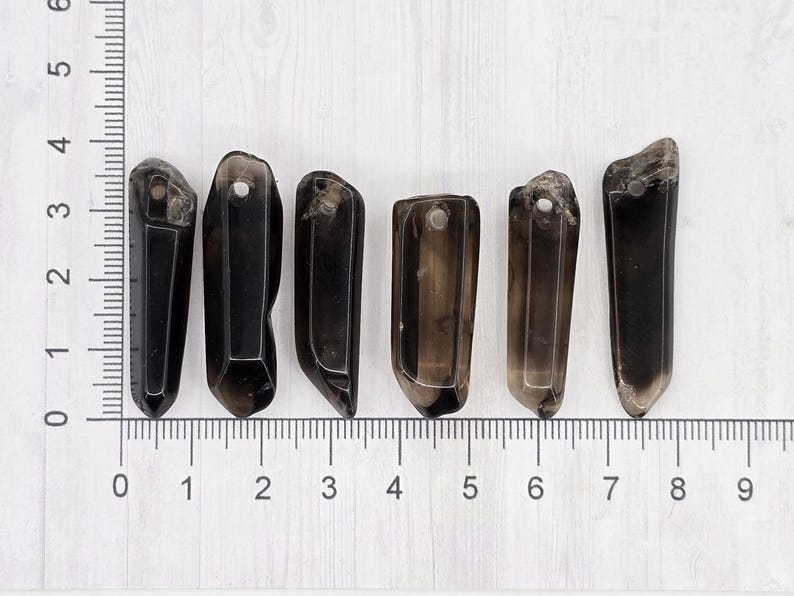 May include: Six dark smoky quartz crystal pendants are arranged on a white surface next to a ruler. Each crystal is a different shape, with a hole drilled at the top for hanging. The crystals range from 1 to 2 inches in length.