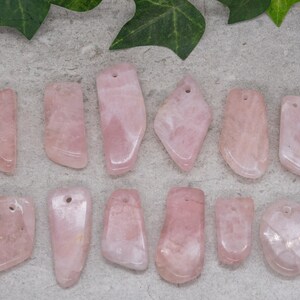 May include: Twelve pink rose quartz pendants with holes drilled at the top. The pendants are various shapes and sizes, with some being more rounded and others more rectangular.