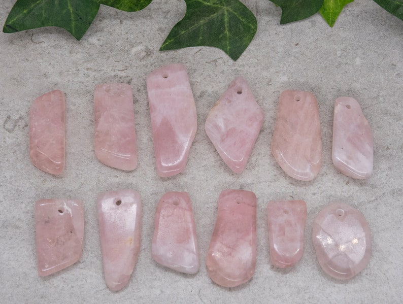 May include: Twelve pink rose quartz pendants with holes drilled at the top. The pendants are various shapes and sizes, with some being more rounded and others more rectangular.