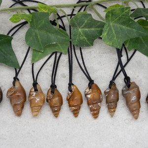 Peut inclure: Dix pendentifs en forme d'escargot en coquille de fossile brune sur des cordons noirs. Les pendentifs sont de formes et de tailles différentes, mais ils sont tous sculptés en forme d'escargot.