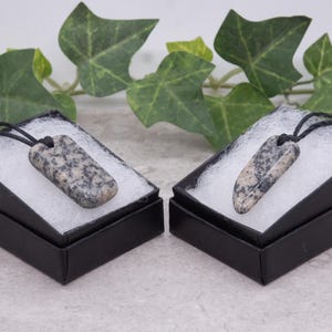 May include: Two black boxes with white interior padding, each containing a grey and black stone pendant on a black cord.