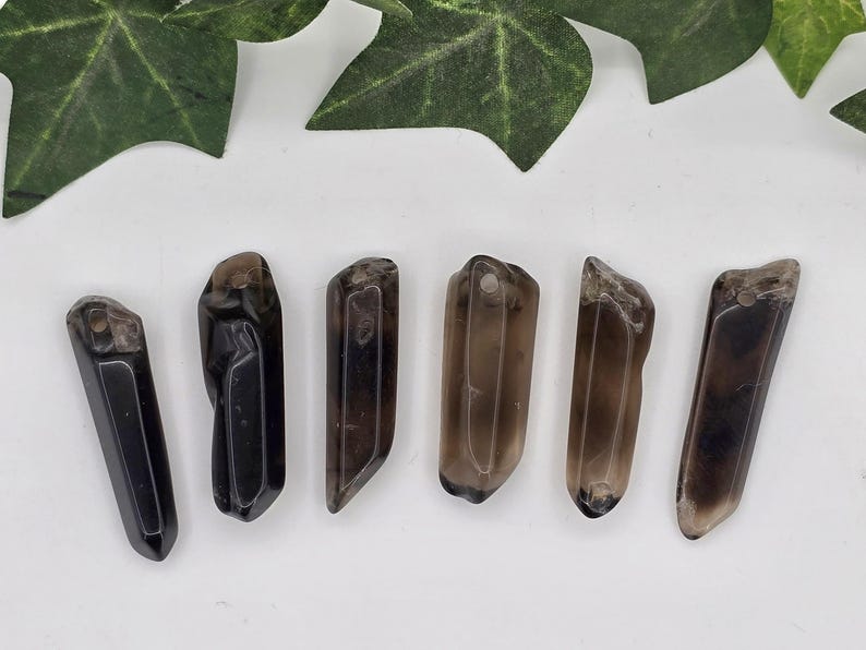 May include: Six polished, elongated smoky quartz crystal pendants. Each pendant is a different shade of brown, ranging from nearly black to light tan. They have a hole drilled at the top for stringing. The crystals are displayed on a white surface with green leaves.