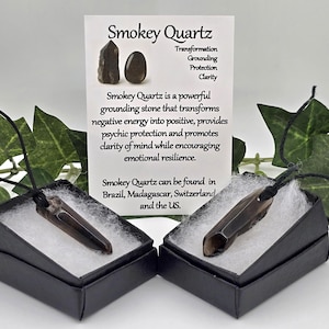 May include: Two Smokey Quartz pendants in black boxes with white filling, displayed with a card detailing the stone's properties. The card reads "Smokey Quartz" and lists benefits like transformation and grounding. Green leaves frame the display.