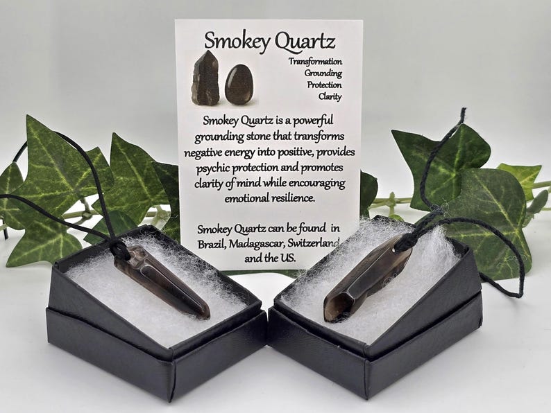 May include: Two Smokey Quartz pendants in black boxes with white filling, displayed with a card detailing the stone's properties. The card reads "Smokey Quartz" and lists benefits like transformation and grounding. Green leaves frame the display.