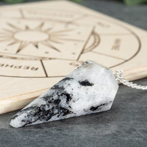 Può includere: Un pendolo di pietra di luna bianca e nera sfaccettata su una catena d'argento. Il cristallo è posto su una tavola di legno con un disegno a raggiera e simboli astrologici. Utilizzato per la divinazione.