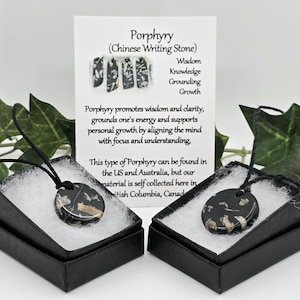 Puede incluir: Dos colgantes de pórfido negro, también conocidos como Piedra de Escritura China, presentados en cajas de regalo negras. Cada colgante es redondo con una base negra y marcas beige. Una tarjeta con texto sobre pórfido está en el fondo.