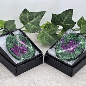 May include: Two green and pink oval pendants in black boxes. The pendants are made of a green stone with pink inclusions. The boxes are lined with white velvet.