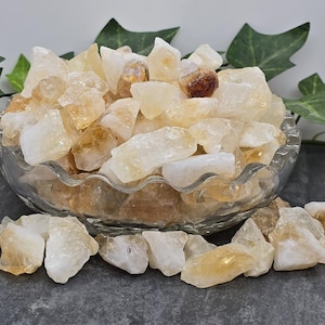 May include: A bowl filled with raw, unpolished, light yellow and orange citrine crystals. The crystals are various sizes and shapes. Some of the crystals are translucent and some are opaque.