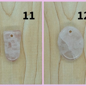 May include: Two pink rose quartz pendants with holes for hanging. The pendant on the left is a freeform shape, and the pendant on the right is oval shaped. Both pendants are on a light brown wooden surface.