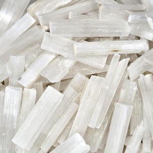 May include: A close-up of a collection of raw, white selenite crystals. The crystals are rectangular and translucent, with a slightly rough texture. They vary in size and are arranged in a random, overlapping pattern.