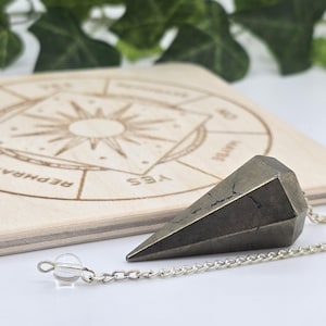 May include: A dark grey faceted pendulum with a silver chain and a clear crystal bead. The pendulum rests on a wooden divination board with a sunburst design and text. The board is light brown.