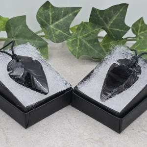 May include: Two black obsidian arrowhead pendants, presented in small black gift boxes with white padding. The pendants are on black cords. Green leaves are in the background, creating a natural setting.