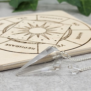 May include: A clear quartz crystal pendulum with a silver chain hanging over a wooden board with a circular chart that says "Yes", "No", "Maybe", "Rephrase", and "Don't Know".