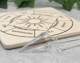 Puede incluir: Un péndulo de cristal de cuarzo transparente con una cadena plateada que cuelga sobre una tabla de madera con un gráfico circular que dice "Sí", "No", "Tal vez", "Reformular" y "No lo sé".