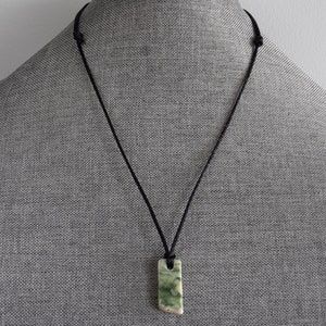 May include: A green and white stone pendant on a black cord necklace. The pendant is a rectangular shape with rounded edges.