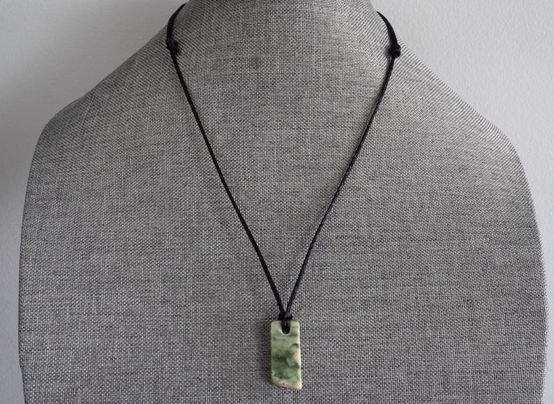 May include: A green and white stone pendant on a black cord necklace. The pendant is a rectangular shape with rounded edges.