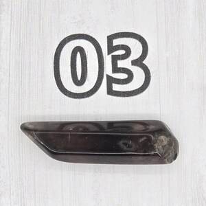 May include: A dark, elongated crystal with a polished surface. The crystal is a deep brown color and has a slightly irregular shape. The number "03" is printed above the crystal in a bold, black font.