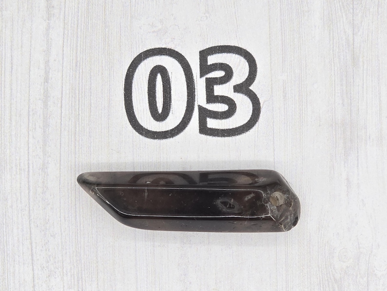 May include: A dark, elongated crystal with a polished surface. The crystal is a deep brown color and has a slightly irregular shape. The number "03" is printed above the crystal in a bold, black font.