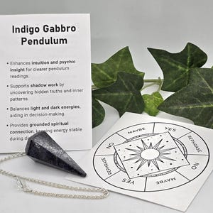 May include: A dark gray, faceted Indigo Gabbro pendulum on a silver-toned chain, accompanied by an informational card titled Indigo Gabbro Pendulum and a circular chart featuring the words Yes, No, Maybe, and Rephrase around a central sun design.