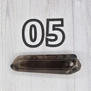 May include: A dark, smoky quartz crystal with a rectangular shape and a hole drilled near one end. The number "05" is printed in black on the white wooden surface behind the crystal.