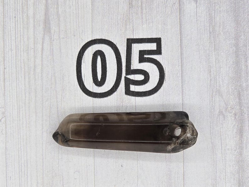 May include: A dark, smoky quartz crystal with a rectangular shape and a hole drilled near one end. The number "05" is printed in black on the white wooden surface behind the crystal.