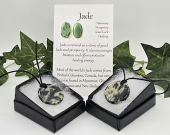 May include: Two jade pendant necklaces in black boxes with white interiors. The round pendants have black cords. A card behind the boxes reads "Jade" and lists its properties, including harmony and prosperity.
