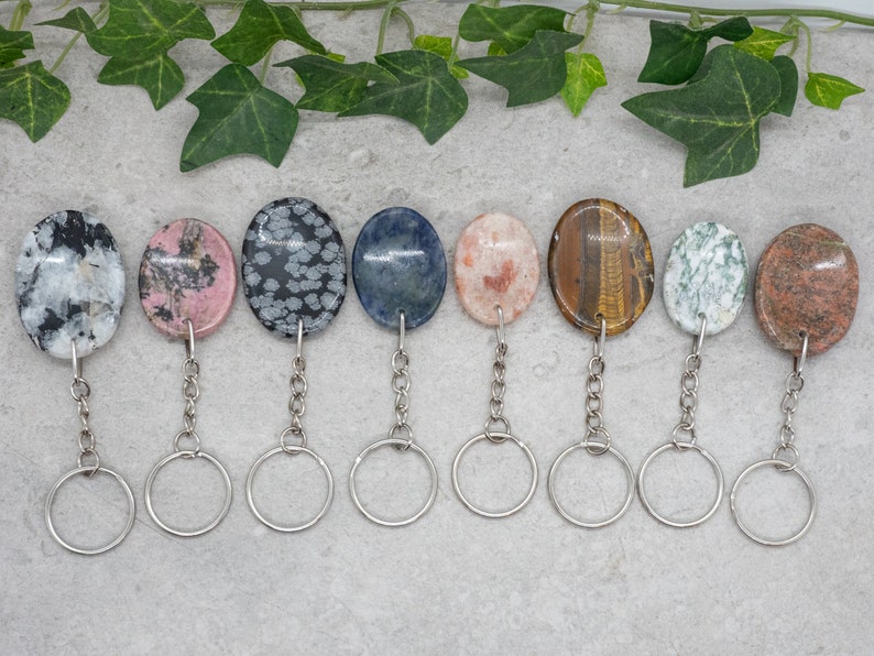 Op de afbeelding: Negen verschillende ovaalvormige edelsteen sleutelhangers met zilveren sleutelringen. De stenen zijn in verschillende kleuren en patronen, waaronder wit, roze, grijs, blauw, oranje, bruin, groen en rood.