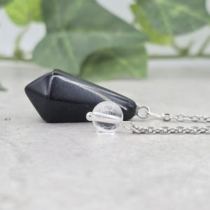 May include: A black crystal pendulum with a silver chain and a clear crystal point. The pendulum is hanging from a silver chain.