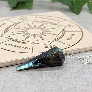 Op de afbeelding: Een zwart en blauw kristallen slinger met een zilveren ketting die hangt boven een houten bord met een cirkelvormig diagram waarop 'Ja', 'Nee', 'Misschien' en 'Hervormuleren' staat.