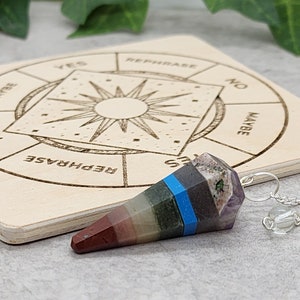 May include: A seven-chakra crystal pendulum with a silver chain and a clear crystal bead. The pendulum is resting on a wooden board with a circular chart that says "Yes", "Rephrase", "No", and "Maybe".