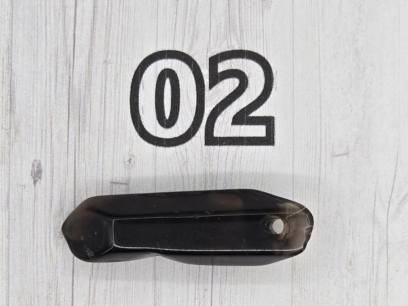 May include: A dark, faceted crystal wand with a small hole at one end. The number "02" is printed in large, bold, black font above the crystal on a light wood-grain background.