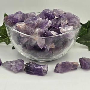 May include: A clear glass bowl filled with raw, jagged amethyst crystals in various shades of purple. Several individual rough amethyst stones are scattered on the surface in front of the bowl, and green ivy leaves are visible on both sides.