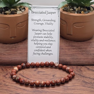 May include: A beaded bracelet made of round, polished brecciated jasper stones in shades of reddish-brown and tan. The bracelet is displayed in front of a white informational card with black text that reads Brecciated Jasper, Strength, Grounding, Courage, Vitality.