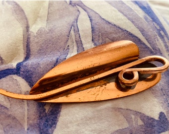May include: A copper-colored leaf-shaped brooch with a curved stem and a looped accent. The brooch has a polished finish and is set against a blue and white patterned fabric. This vintage-style accessory is suitable for adding a touch of nature-inspired elegance.
