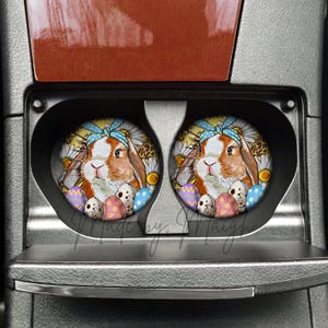 May include: Two round car coasters featuring a brown and white bunny wearing a blue bow, surrounded by Easter eggs and daisies. The coasters are set in a car's cup holders.