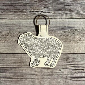 May include: White leather keychain with a black outline of a sheep. The sheep is filled with a maze pattern.