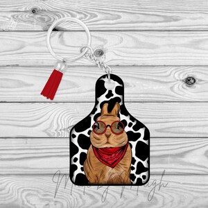 May include: A novelty keychain featuring a cartoon rabbit wearing red glasses and a red bandana. The rabbit is on a black and white cow print tag, attached to a silver chain with a red tassel.