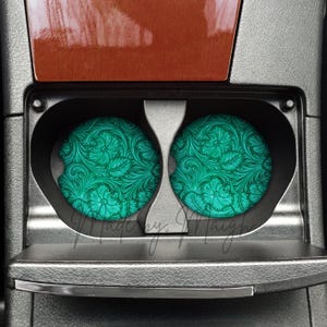 May include: Two teal car coasters with a floral pattern are placed in a car's cup holders. The coasters have a detailed design of flowers and leaves. The car's interior features a brown wood-like trim and a gray console.