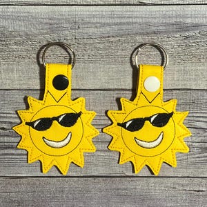 May include: Two yellow sun-shaped keychains with black sunglasses and smiling faces. Each keychain has a yellow strap with a metal ring and a black or white button. The keychains are on a wooden background.