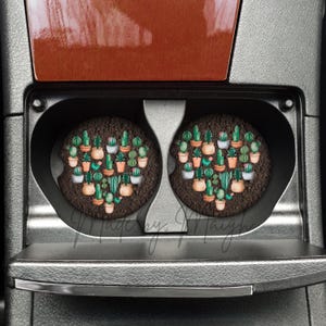 May include: Two car coasters with a heart-shaped design of potted cacti and succulents. The coasters are round, with a dark brown background, and are placed in a car's cup holders. The image is taken from a top-down perspective.