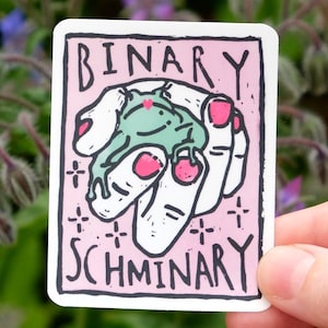 May include: A rectangular sticker with a light pink background featuring a black-outlined illustration of a hand holding a small green frog. The text "BINARY SCHMINARY" is printed in black capital letters above and below the central illustration.