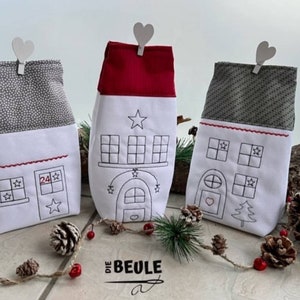 May include: Three white fabric advent calendar houses with red and gray accents. Each house has a different design with embroidered details, including windows, stars, a heart, and a Christmas tree. The houses are decorated with white hearts and are sitting on a wooden surface.