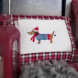 May include: A white pillow with a red and black plaid border features an embroidered image of a dachshund wearing a Santa hat, a blue sweater, and a red scarf.