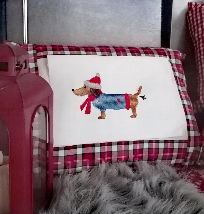 May include: A white pillow with a red and black plaid border features an embroidered image of a dachshund wearing a Santa hat, a blue sweater, and a red scarf.