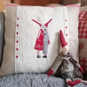 Könnte beinhalten: Ein weißes Leinenkissen mit rotem Besatz und einem roten und grauen Stickmuster von zwei Figuren, die rote Hüte tragen. Eine rote und graue Gnompuppe sitzt auf dem Kissen.