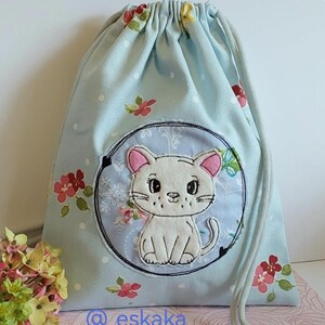 Könnte beinhalten: Hellblaues Kordelzugbeutel mit Blumenmuster und einem weißen Katzen-Applikation. Die Katze hat rosa Ohren und ist in einem schwarz umrandeten Kreis zentriert. Der Beutel hat einen hellblauen Kordelzug und der Text "@_eskaka_" ist sichtbar.