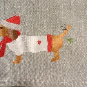 May include: Embroidered dachshund wearing a red and white Santa hat and scarf, holding a sprig of holly in its mouth. The dachshund is embroidered on a gray linen fabric with the text "Handmade with Love" in a brown circle.