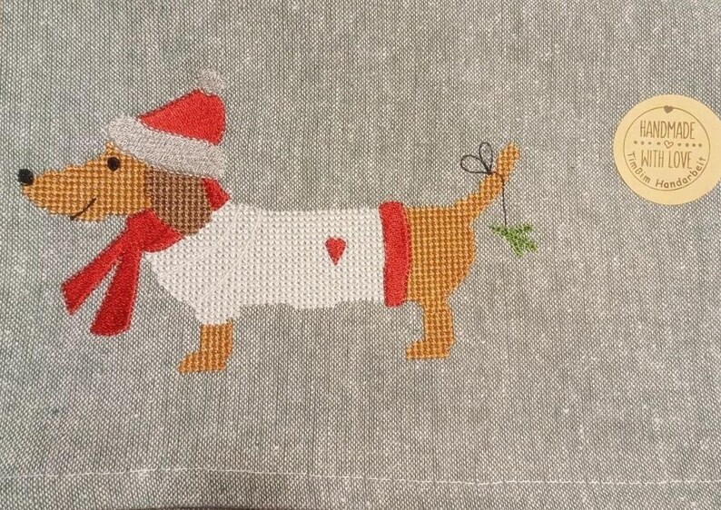 May include: Embroidered dachshund wearing a red and white Santa hat and scarf, holding a sprig of holly in its mouth. The dachshund is embroidered on a gray linen fabric with the text "Handmade with Love" in a brown circle.