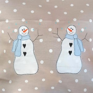 Könnte beinhalten: Zwei weiße Filz-Schneemann-Applikationen mit schwarzen Knöpfen und blauen Schals auf einem beigefarbenen Hintergrund mit weißen Punkten.