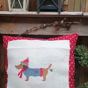 May include: A white pillow with a red polka dot border features a cross-stitch design of a dachshund wearing a blue sweater, red scarf, and a red hat. The dachshund is holding a sprig of holly in its mouth.